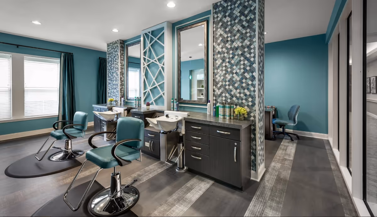 Interior view of a modern salon area with teal walls and two teal salon chairs in front of wash basins and large mirrors. The space features patterned tile columns, dark cabinetry with hair care products on the counter, and a small desk with a chair in the background.