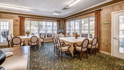Dining room with several round tables covered in tablecloths, upholstered chairs, floral centerpieces, and large windows letting in daylight.