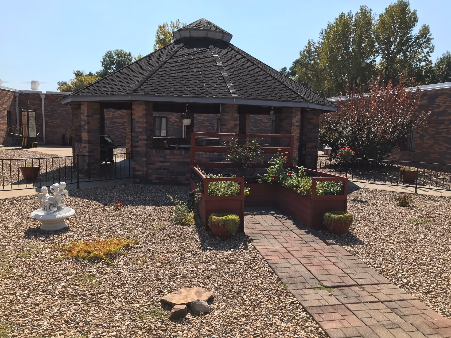 Outdoor courtyard area with a brick gazebo structure in the center, surrounded by gravel ground cover, potted plants, and a small garden bed with flowers. There is a paved walkway leading to the gazebo, and trees and shrubs are visible in the background under a clear blue sky.