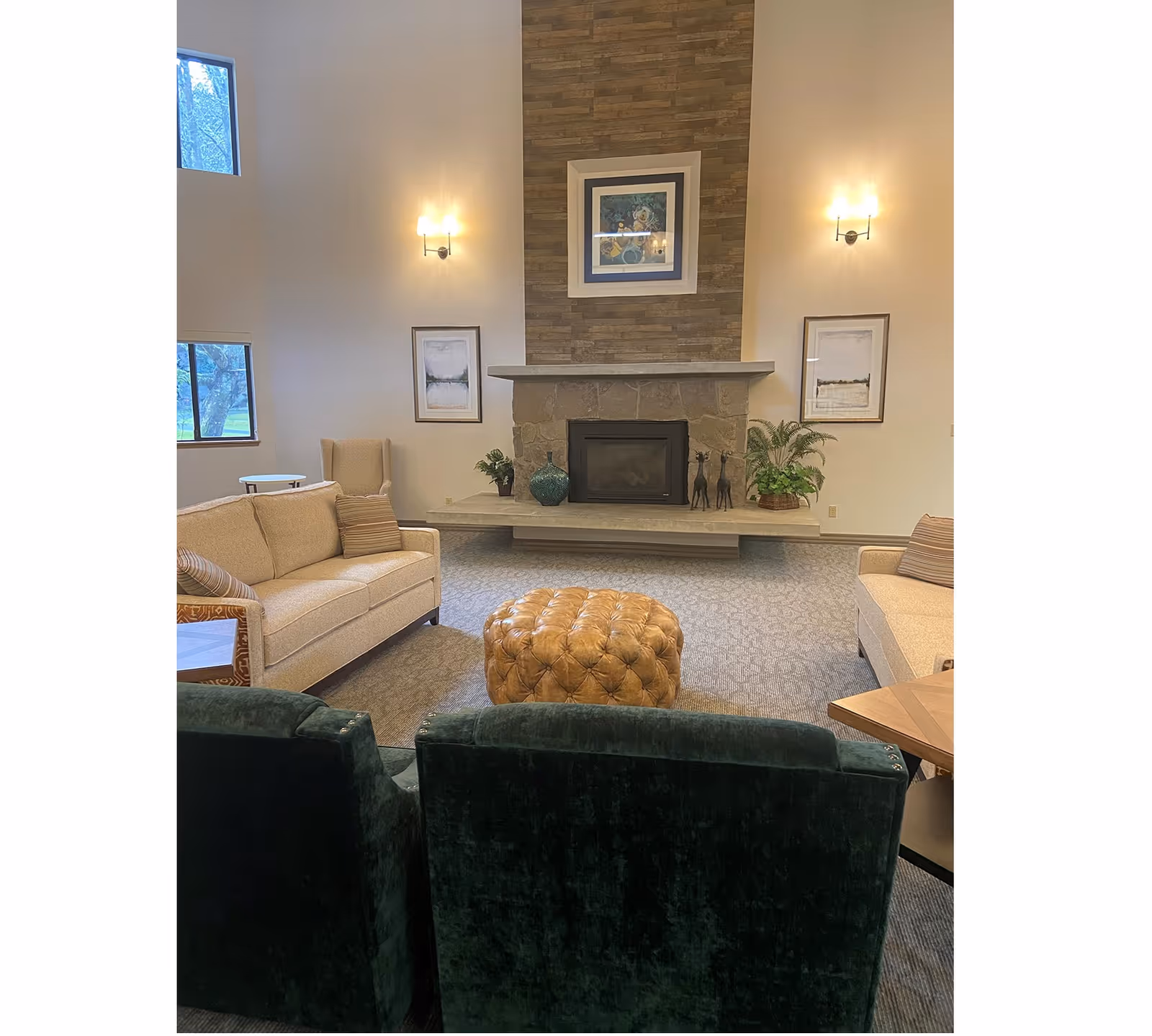 Cozy senior living common room with sofas and chairs arranged around a tufted ottoman facing a stone fireplace with artwork and plants.