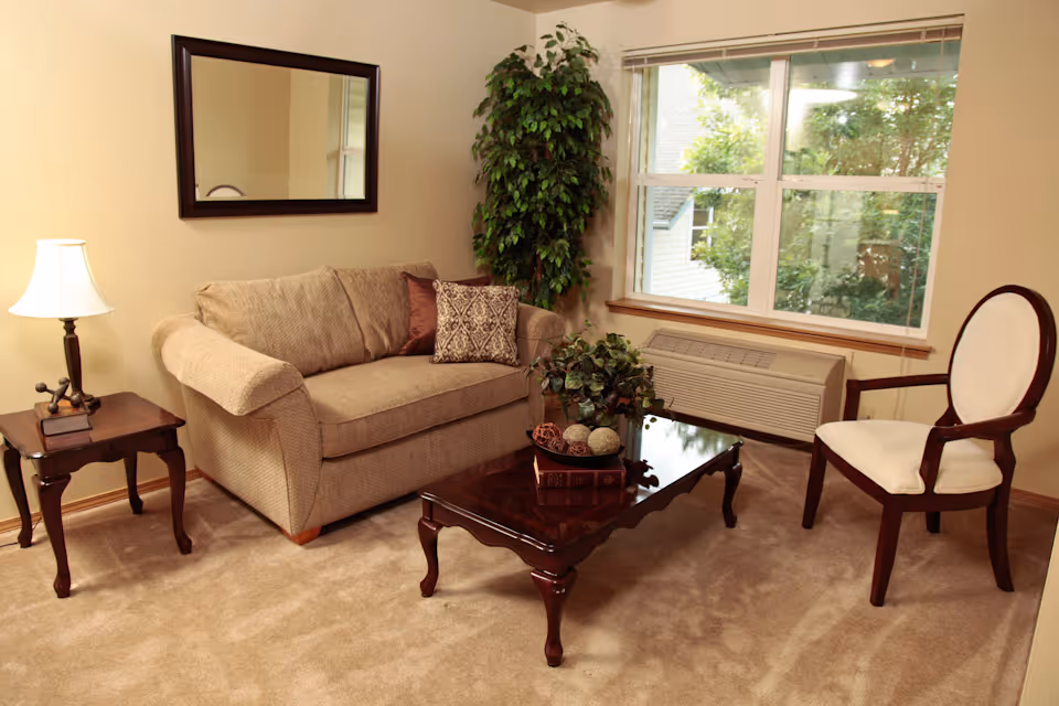 A cozy living room with a beige loveseat adorned with two decorative pillows, a wooden coffee table with decorative items, a wooden side table with a lamp and books, a white cushioned wooden chair, a large green potted plant in the corner, a window showing greenery outside, and a wall-mounted mirror above the loveseat.