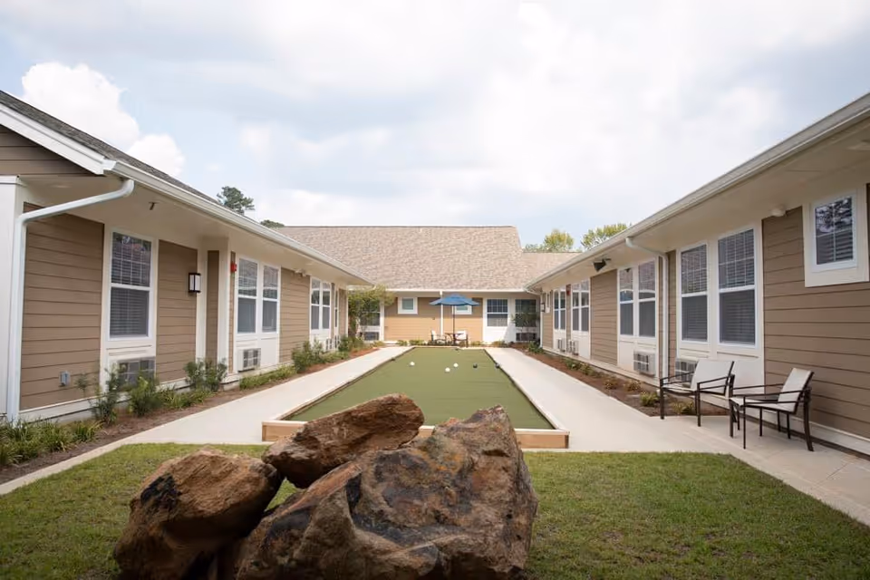 Outdoor courtyard area at New Perspective Senior Living | West Fargo featuring a bocce ball court in the center, surrounded by two single-story buildings with beige siding and white trim. There are chairs and small tables along the walkways, and large rocks in the foreground with a partly cloudy sky above.