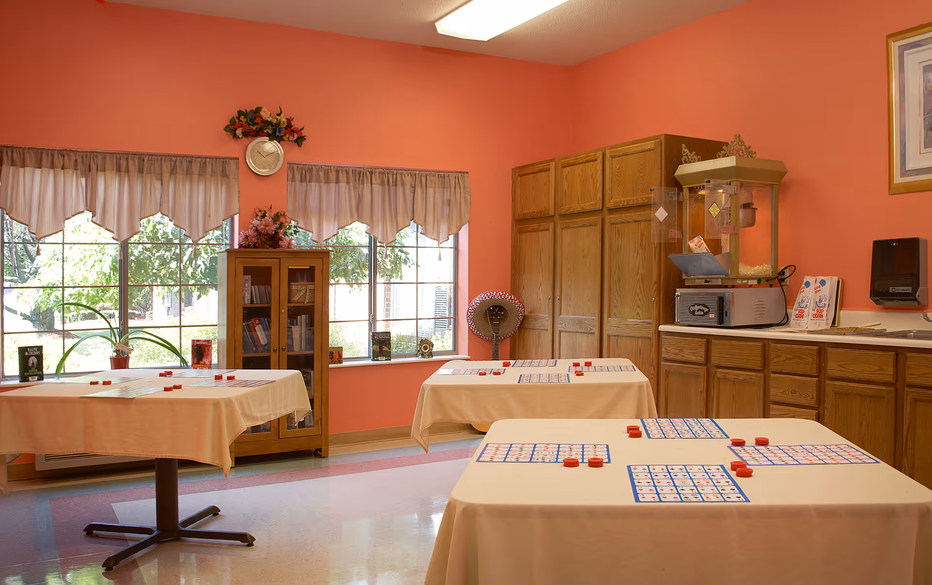 Bright activity room with tables covered in bingo cards and markers, wood cabinets, a popcorn machine, and large windows.
