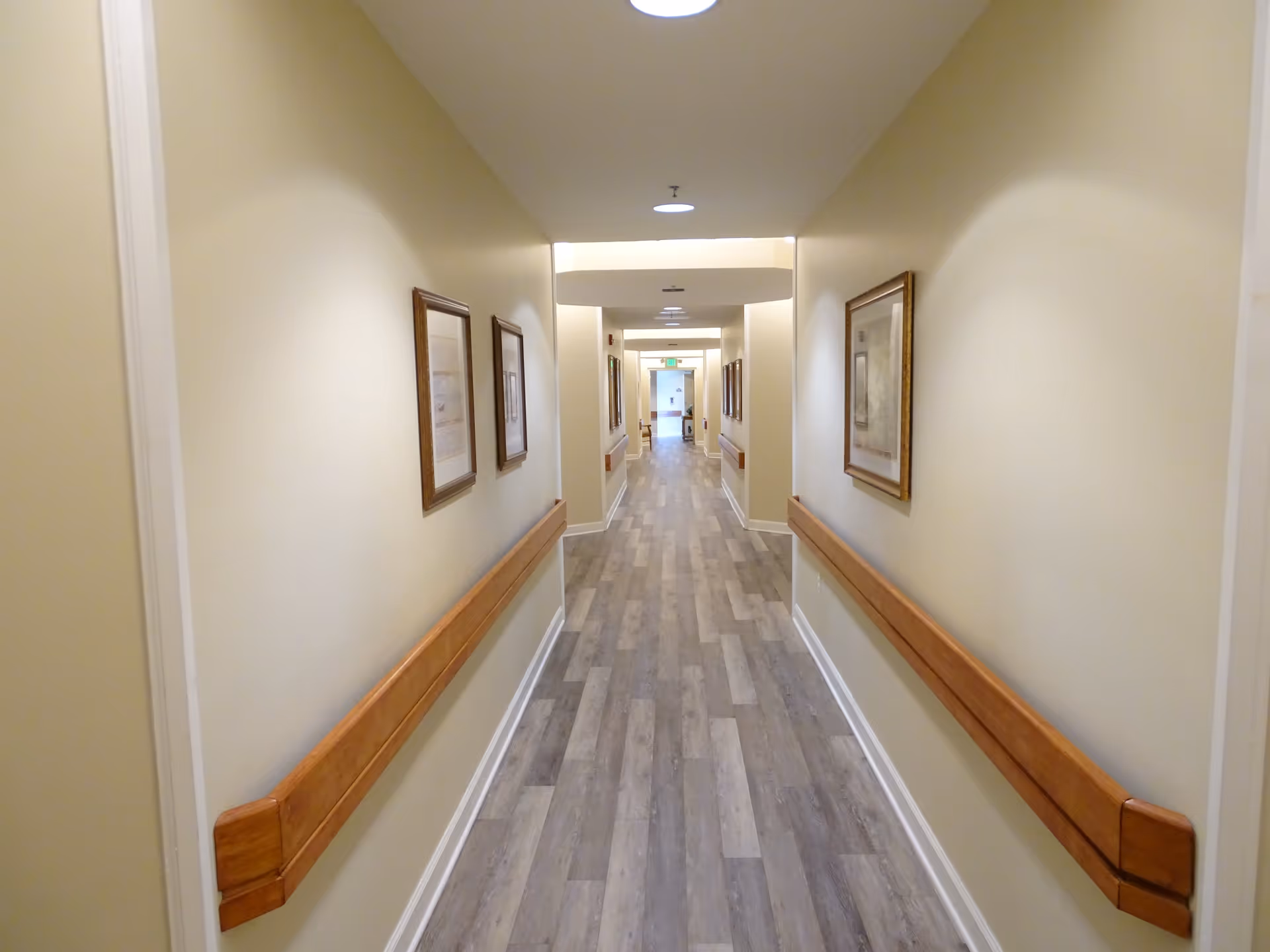 A long, well-lit hallway in a senior living facility with light beige walls, wooden handrails on both sides, framed artwork hanging on the walls, and a wood-patterned floor leading to a door at the far end.