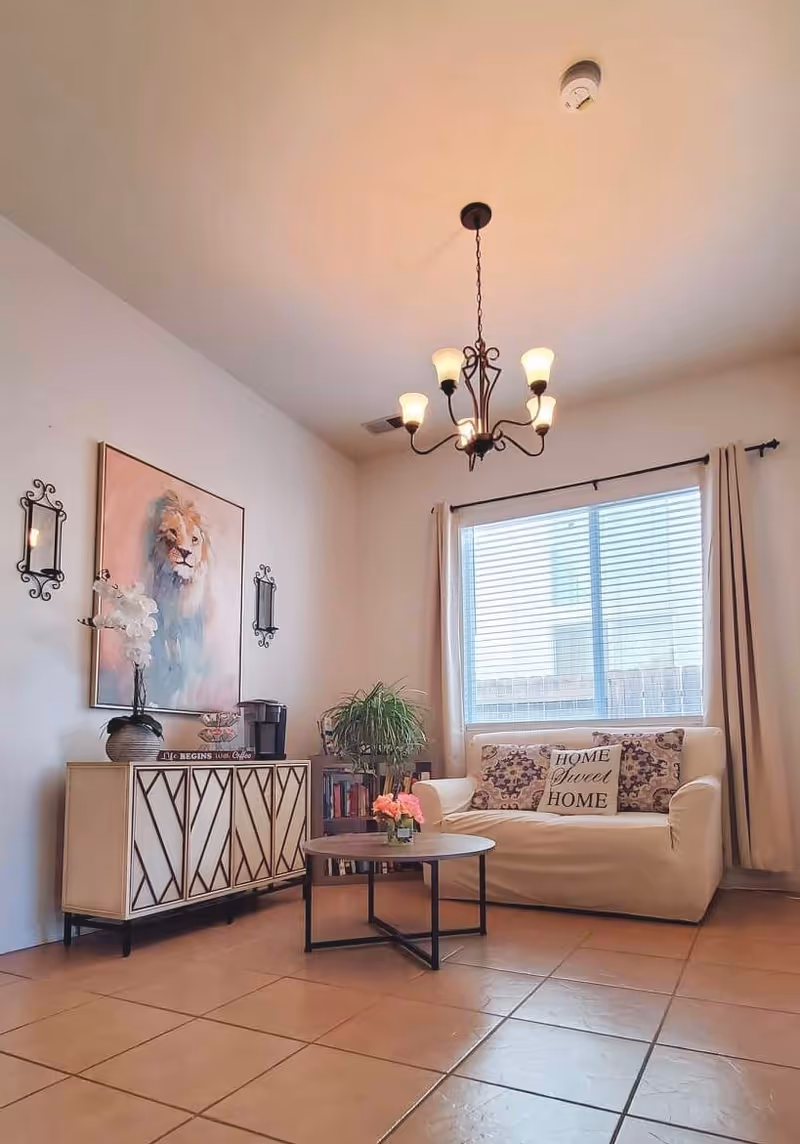 A cozy living room with a white loveseat adorned with three decorative pillows, one of which says 'HOME Sweet HOME'. A round coffee table with a small flower arrangement sits in front of the loveseat. To the left, there is a stylish cabinet with a vase of white flowers, a coffee maker, and some books. Above the cabinet hangs a large painting of a lion flanked by two decorative wall sconces. A large window with beige curtains lets in natural light, and a chandelier with six lights hangs from the ceiling. The floor is tiled in a warm brown color.
