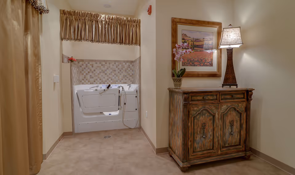 A walk-in bathtub with a tiled wall surround and a handheld showerhead is situated in a small room. To the right, there is a rustic wooden cabinet with drawers and doors, topped with a table lamp and a potted orchid. Above the cabinet hangs a framed landscape painting. The room has beige walls and floor tiles, with a gold curtain partially visible on the left.