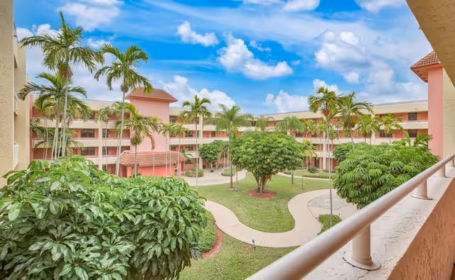 View of a courtyard in Elison Independent Living of Lake Worth featuring a winding paved walkway, green grass, palm trees, and a multi-story building with balconies under a partly cloudy blue sky.