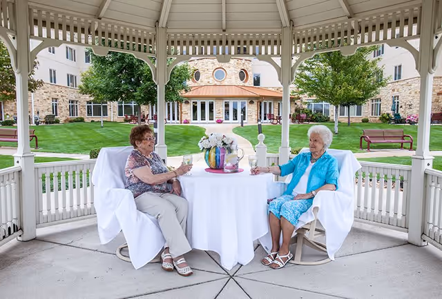 Two elderly women sitting at a round table covered with a white tablecloth under a gazebo. They are outdoors in a courtyard with green grass, trees, and a building with stone walls and windows in the background. The women are holding glasses and appear to be enjoying a conversation.