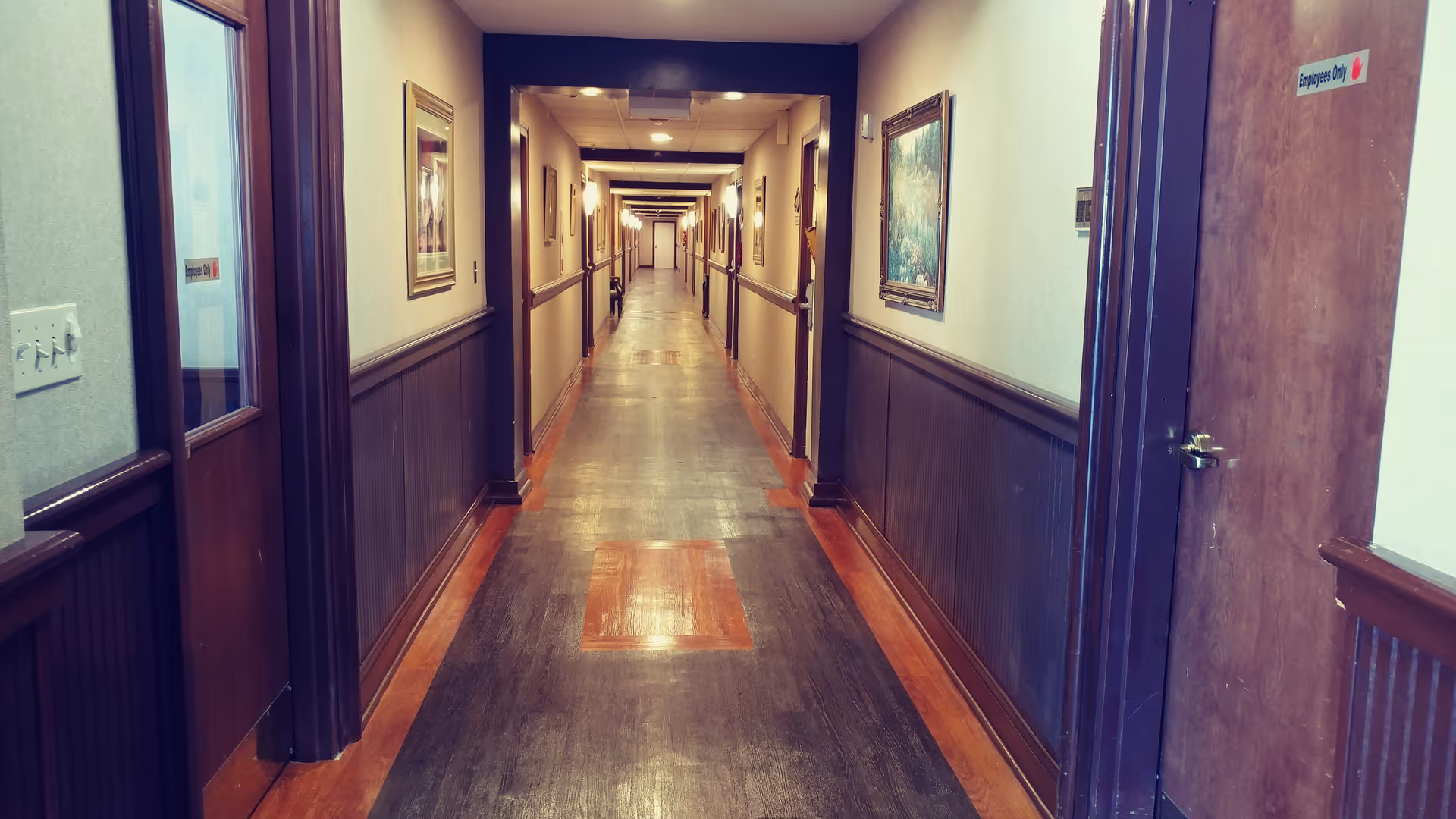 A long, narrow hallway in a senior living facility with wooden paneling on the lower half of the walls and framed paintings hanging on the upper half. The floor is wooden with a polished finish, and several doors line both sides of the corridor. One door on the right has a sign that reads 'Employees Only'.
