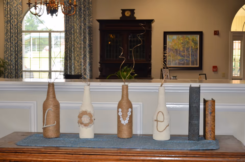 Decorative bottles wrapped in twine and fabric arranged on a wooden table with a blue runner, spelling out the word 'LOVE' with buttons, pearls, and twine. In the background, there is a window with patterned curtains, a chandelier, a dark wooden cabinet, a framed painting, and a doorway.