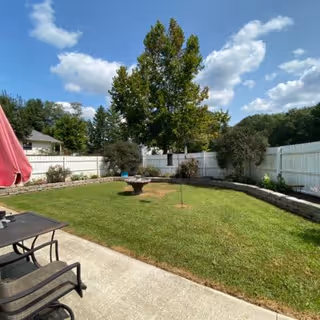 Outdoor garden area with green grass, a birdbath in the center, surrounded by a white fence and trees under a partly cloudy blue sky. There are patio chairs and a table with a red umbrella on a concrete surface in the foreground.