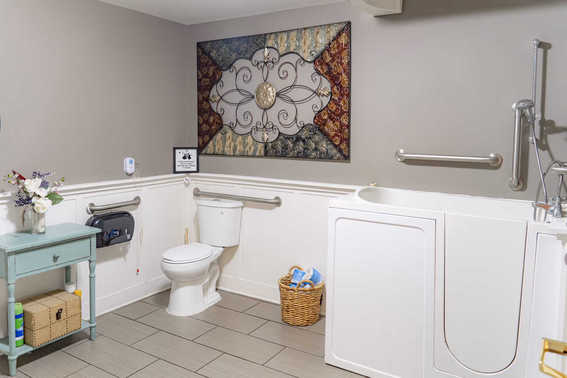 An accessible bathroom featuring a walk-in tub, a toilet, grab bars, and decorative wall art. A small blue table holds a vase of flowers, and a basket with toiletries is on the floor.