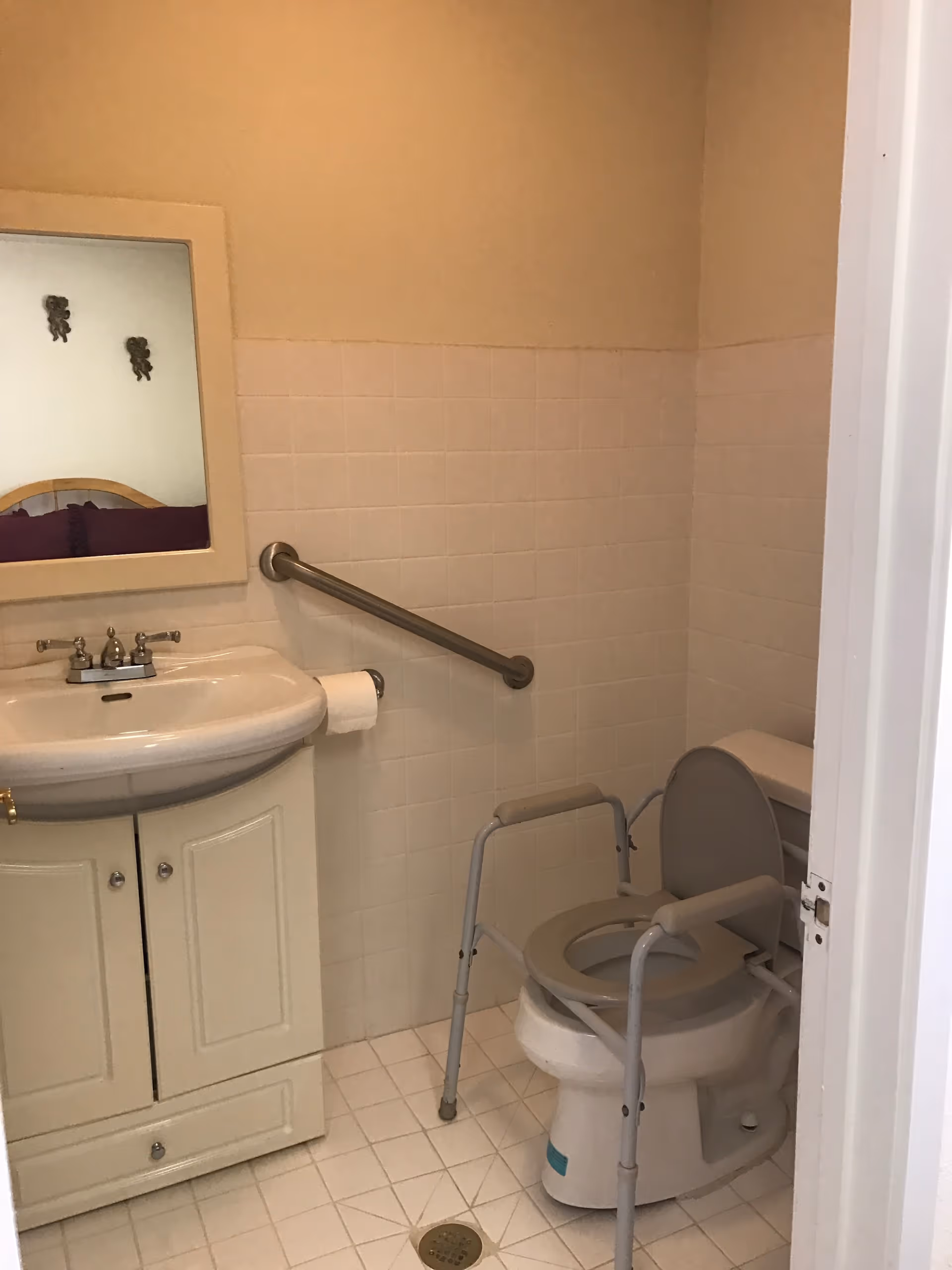 A small bathroom with a white sink and cabinet, a mirror above the sink, a toilet with an elevated seat and arm supports, a grab bar on the wall, and tiled walls and floor.