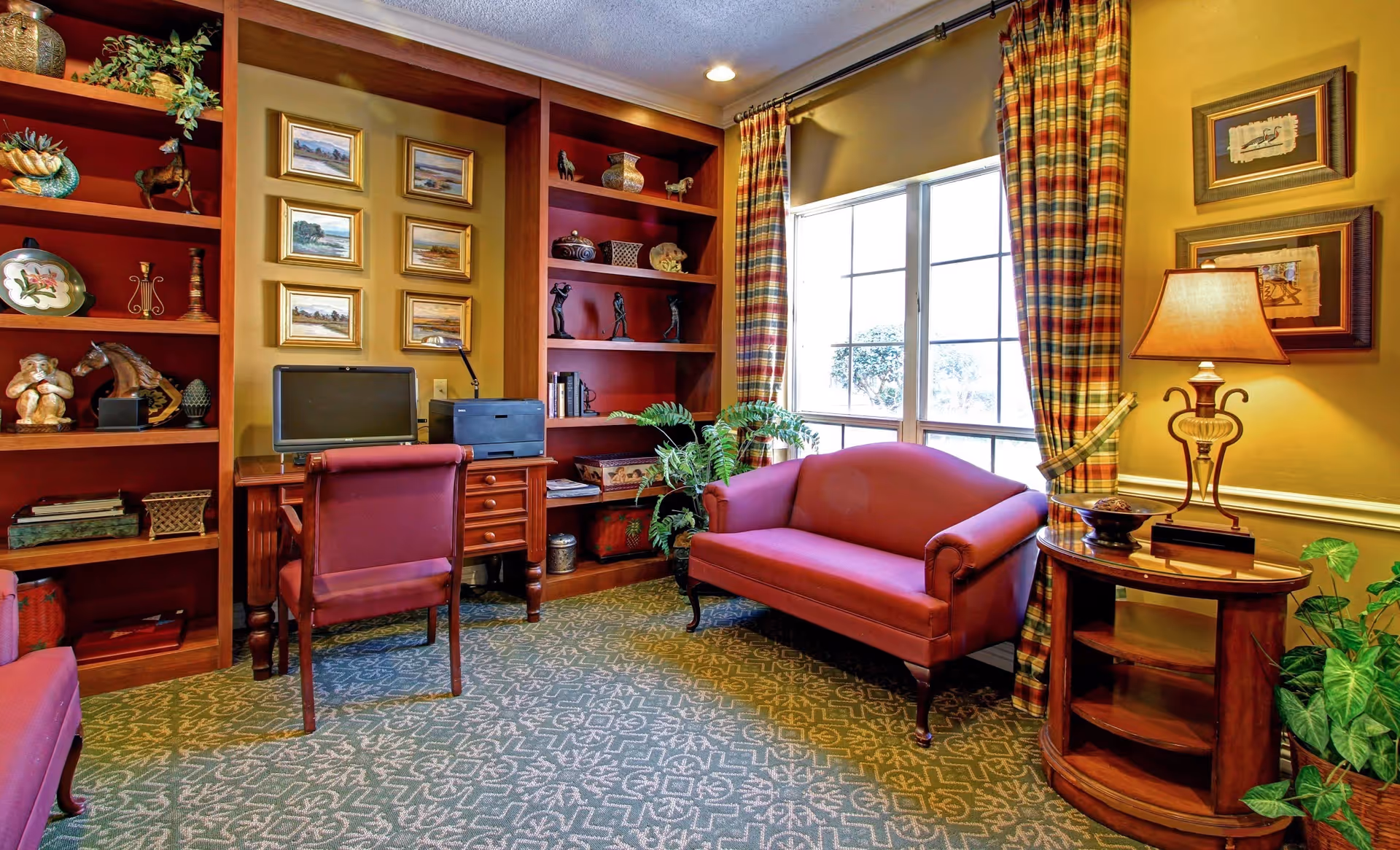 Cozy sitting area with a red upholstered loveseat and chair, wooden built-in bookshelves and a desk beside a large window with plaid curtains.