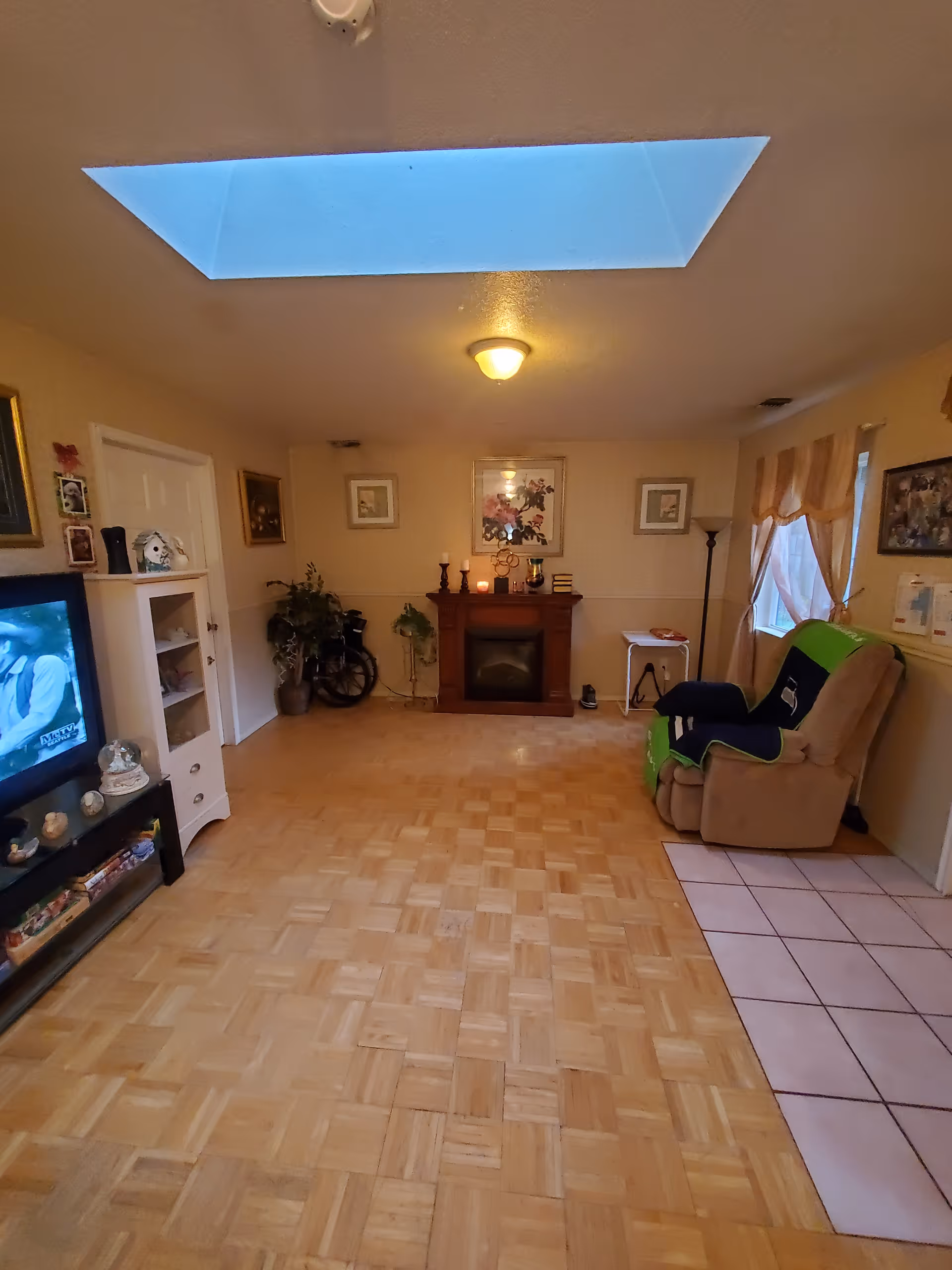 A cozy living room with a wooden parquet floor and a tiled area near a beige recliner chair covered with a green and blue blanket. The room features a fireplace with decorative items and framed pictures on the wall above it. There is a TV on a stand to the left, a white shelving unit, a potted plant, a wheelchair, and a window with beige curtains on the right side. A ceiling light and a skylight provide illumination.