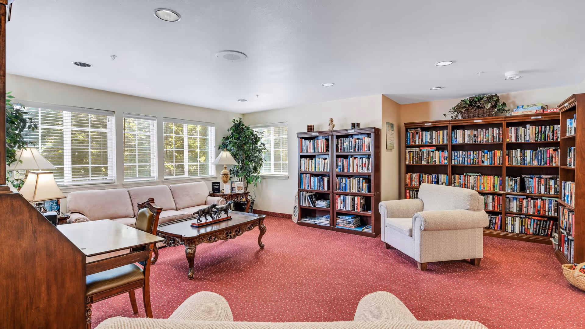 A cozy reading room with large windows letting in natural light, a beige sofa, a wooden coffee table with decorative figurines, a beige armchair, a wooden desk with a chair, and multiple bookshelves filled with books. The room has red carpet flooring and potted plants near the windows.