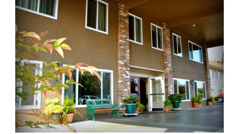 Exterior view of a two-story building with brown walls and brick columns. Several windows are visible on both floors. There is a green bench and multiple potted plants near the entrance, which has glass double doors. A car is partially visible on the right side of the image.