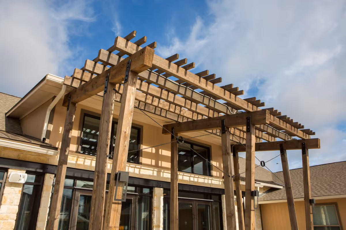 Exterior view of a senior living facility building with a wooden pergola structure in front of large windows under a partly cloudy blue sky.