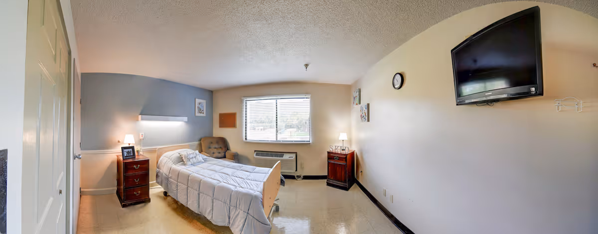 A simple, clean bedroom in a senior living facility with a single bed covered in a white comforter, a wooden nightstand with a lamp and picture frame, a cushioned chair near a window with blinds, a wall-mounted TV, a clock on the wall, and another small wooden cabinet with a lamp.