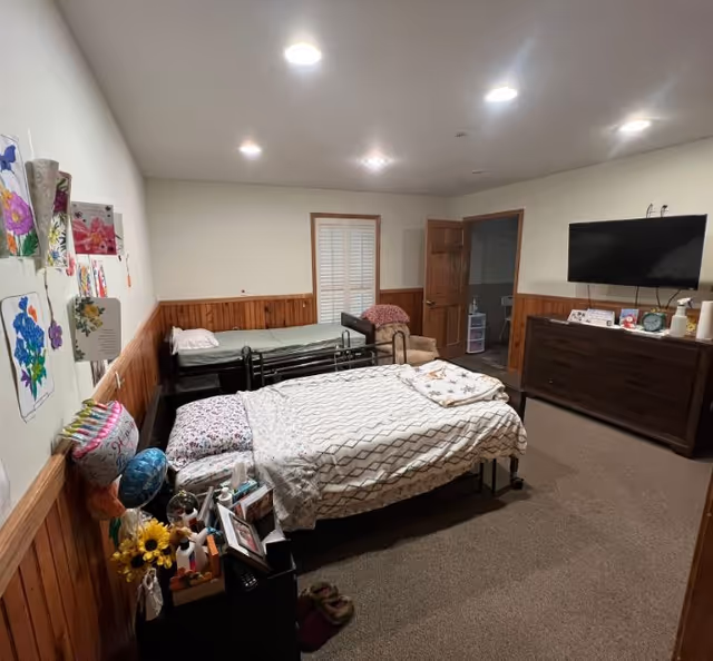 A cozy bedroom in an assisted living facility featuring two beds with patterned bedding. The room has wood paneling on the lower half of the walls and white paint above. There are colorful drawings and cards displayed on one wall. A dresser with a flat-screen TV mounted above it is on the right side of the room. The room is softly lit with recessed ceiling lights, and there is a doorway leading to another room.