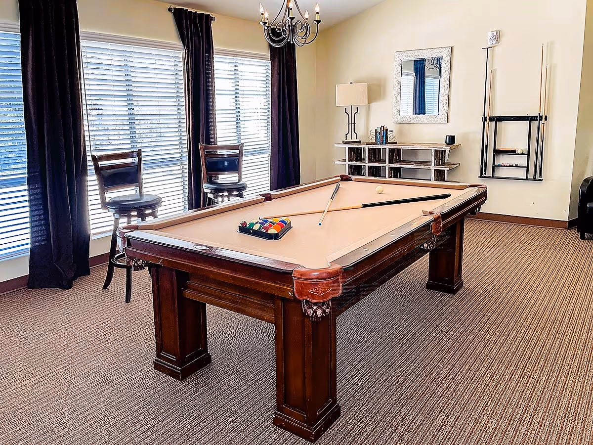 A well-lit game room with a beige pool table in the center, two pool cues resting on the table, a rack of billiard balls, two wooden bar stools by large windows with blinds and dark curtains, a chandelier overhead, a small shelf with books and decor, a wall-mounted mirror, and a wall rack holding additional pool cues.