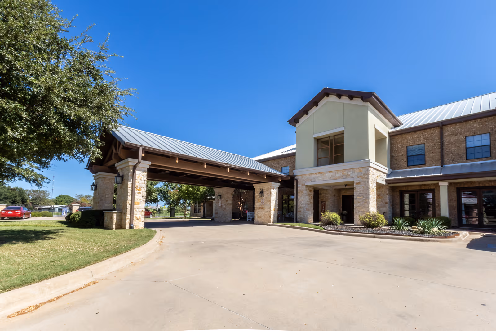 Front entrance of a two-story assisted living building with a covered porte-cochere, stone facade, and driveway.