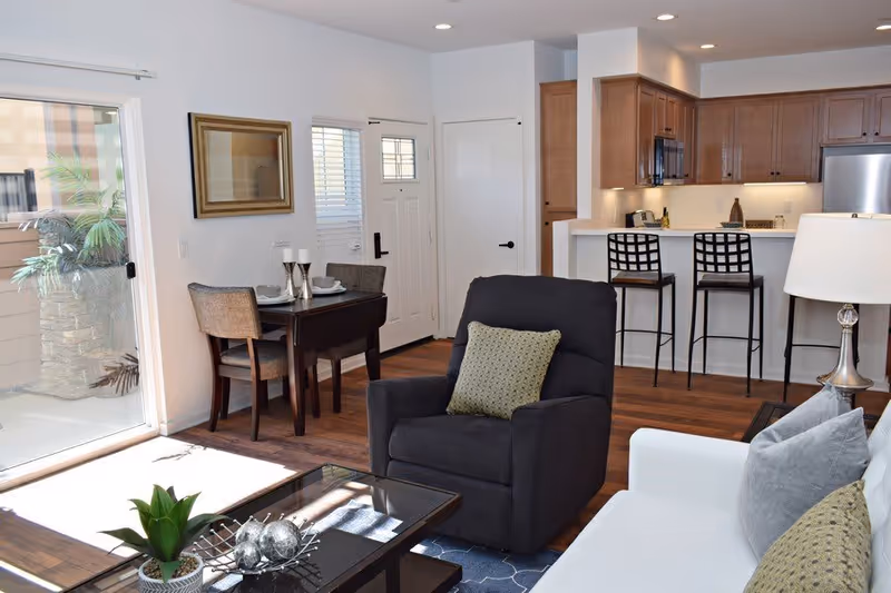 Interior view of a modern living space in an active living community, featuring a black armchair with a green pillow, a white sofa with gray and green pillows, a glass coffee table with decorative items, a small dining table set for two near a sliding glass door, and a kitchen area with wooden cabinets, a breakfast bar with two black bar stools, and stainless steel appliances.