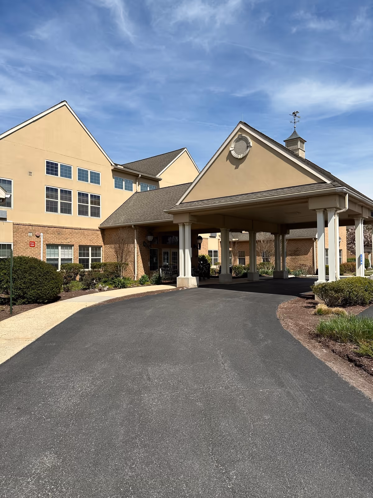 Exterior view of a senior living facility with a covered entrance driveway, beige and brick building facade, and landscaped bushes under a partly cloudy blue sky.