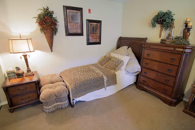 A cozy bedroom with a single bed covered in patterned bedding and pillows. Next to the bed is a wooden nightstand with a lamp, clock, and decorative items. At the foot of the bed are two cushioned ottomans. On the right side, there is a tall wooden dresser with decorative plants and candles on top. The walls are light-colored and adorned with framed artwork and a wall-mounted plant holder.