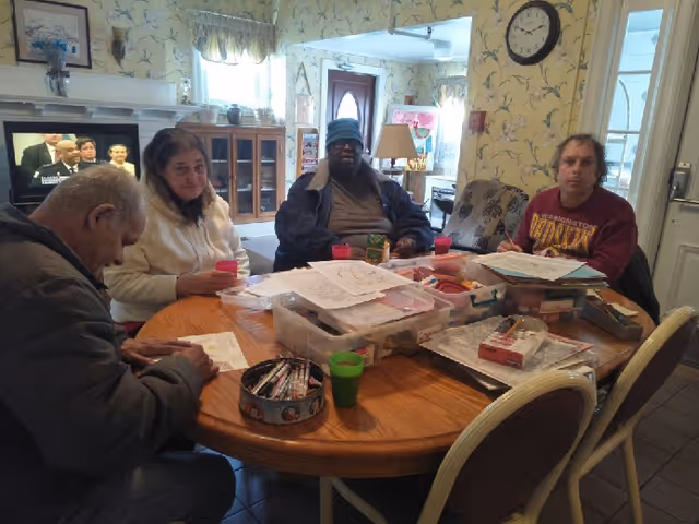 Four people sit around a round table doing crafts and paperwork in a communal room with a TV, shelving, and a door in the background.