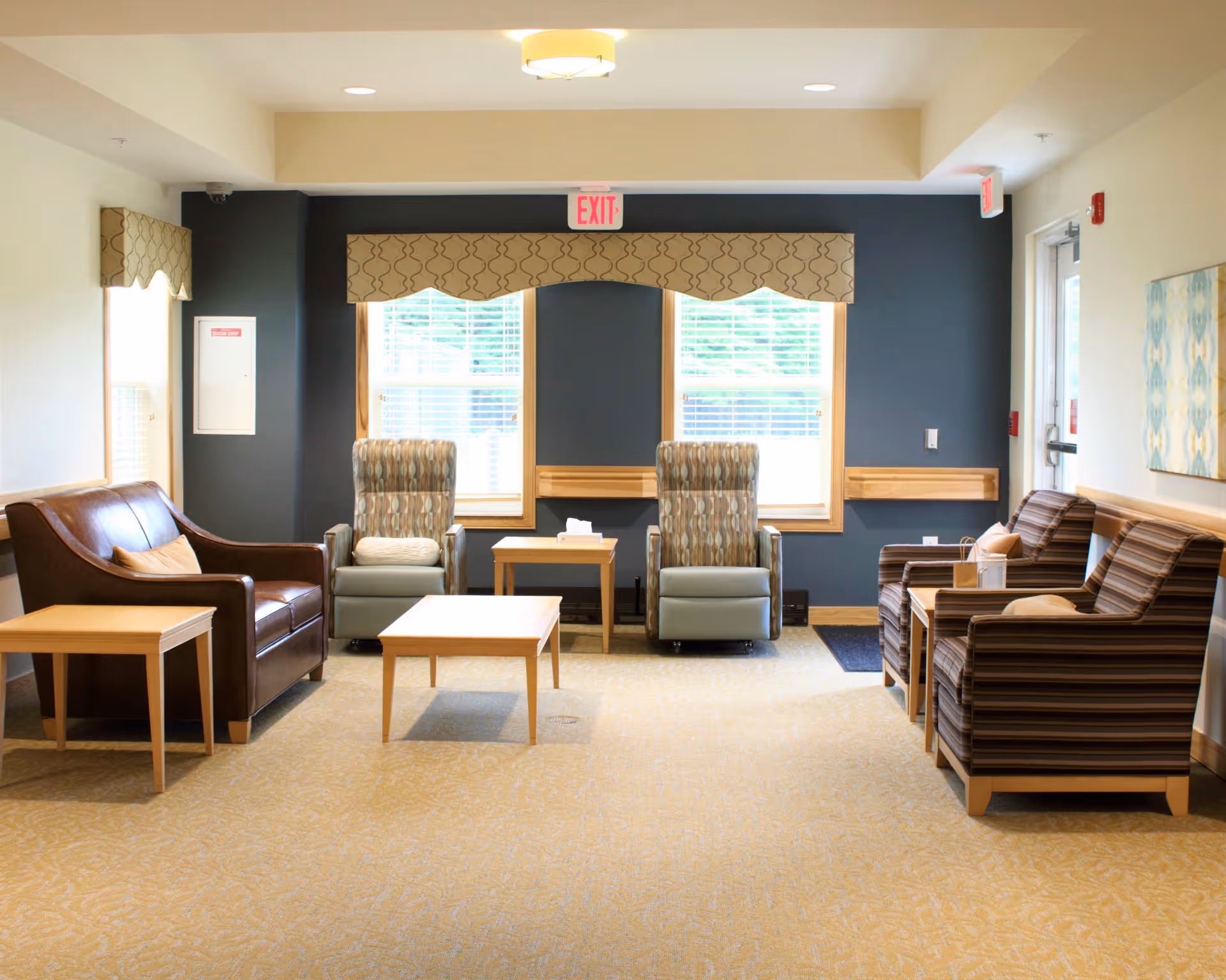 A well-lit common area with a variety of seating including a brown leather loveseat, two patterned armchairs, and two striped armchairs. There are wooden side tables and a coffee table in the center. Two large windows with patterned valances allow natural light to fill the room. The walls are painted in a combination of beige and dark blue, and there is an exit sign above the windows.
