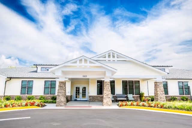 Front exterior view of Chantilly Heights Memory Care facility with a covered entrance supported by stone pillars, large windows, and landscaped flower beds under a partly cloudy blue sky.