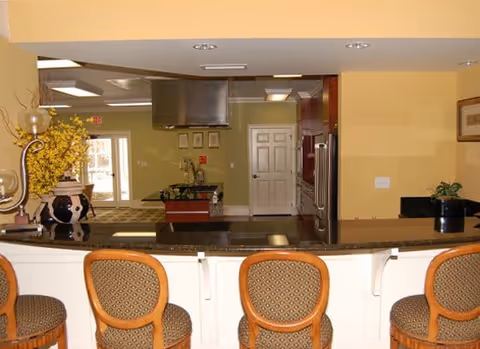 Interior view of a communal kitchen/bar area with four upholstered bar stools at a granite counter facing an open kitchen.