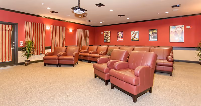 A spacious lounge area with red leather armchairs and sofas arranged in rows facing forward. The walls are painted red with several framed vintage movie posters hanging. There are striped curtains on the windows and two potted plants near the door and corner. The room has beige carpet flooring and ceiling lights.