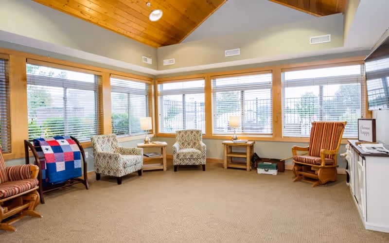 A bright and cozy living room area with large windows featuring wooden frames and blinds. The room has a high wooden ceiling and beige carpet. There are four upholstered chairs arranged around two wooden side tables with lamps. A colorful patchwork quilt is draped over a wooden rack. A flat-screen TV is mounted on the wall above a white cabinet with a small sign that reads 'choose happy.'