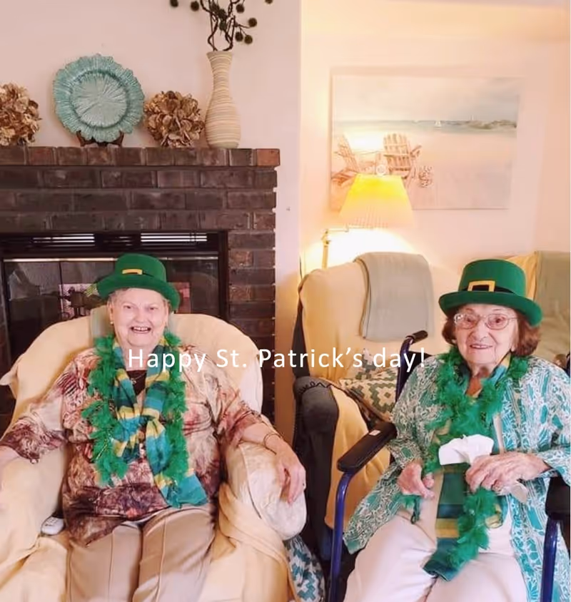 Two elderly women sitting indoors in a living room setting, both wearing green St. Patrick's Day hats and green feather boas. One woman is seated in a beige armchair in front of a brick fireplace decorated with a green plate and dried flowers. The other woman is seated in a wheelchair next to a beige armchair with a lamp and a beach-themed painting on the wall behind them. Text on the image reads 'Happy St. Patrick's day!'