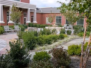 Outdoor courtyard area with paved walkways, green shrubs, small trees, and several chairs and benches arranged for seating near a red brick building.