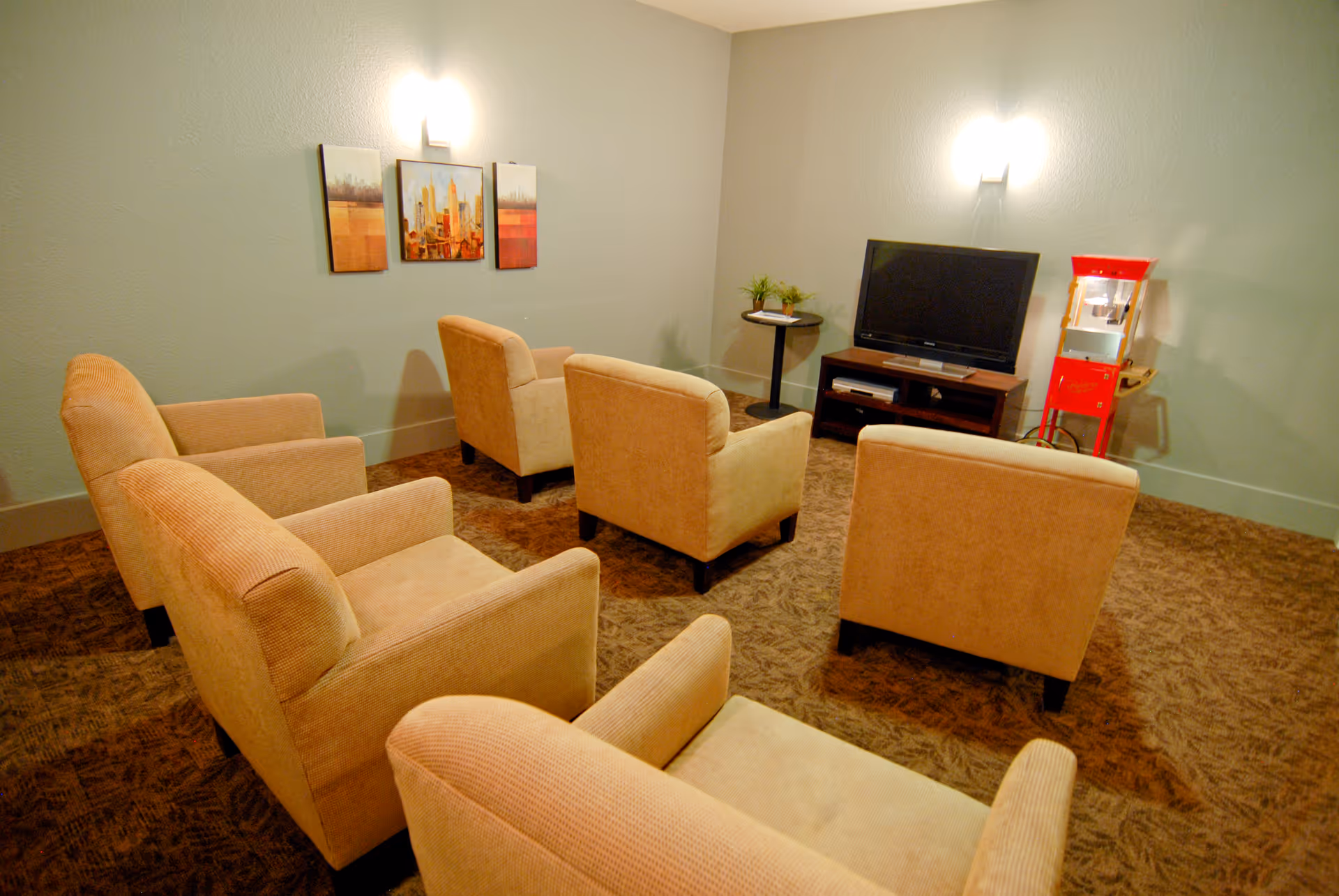 A cozy living room with six beige armchairs arranged in two rows facing a flat-screen TV on a wooden stand. On the right side, there is a red popcorn machine. The walls are light green with two wall-mounted lights and three framed pictures hanging above the chairs.
