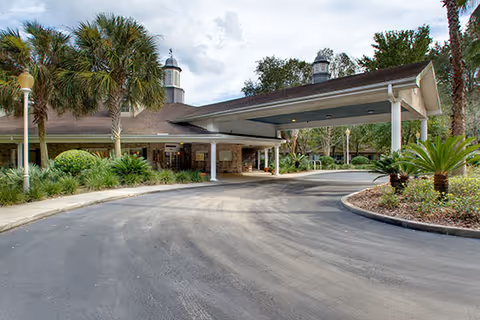 Covered porte-cochère entrance and circular paved driveway in front of a single-story building surrounded by palm trees and landscaping.