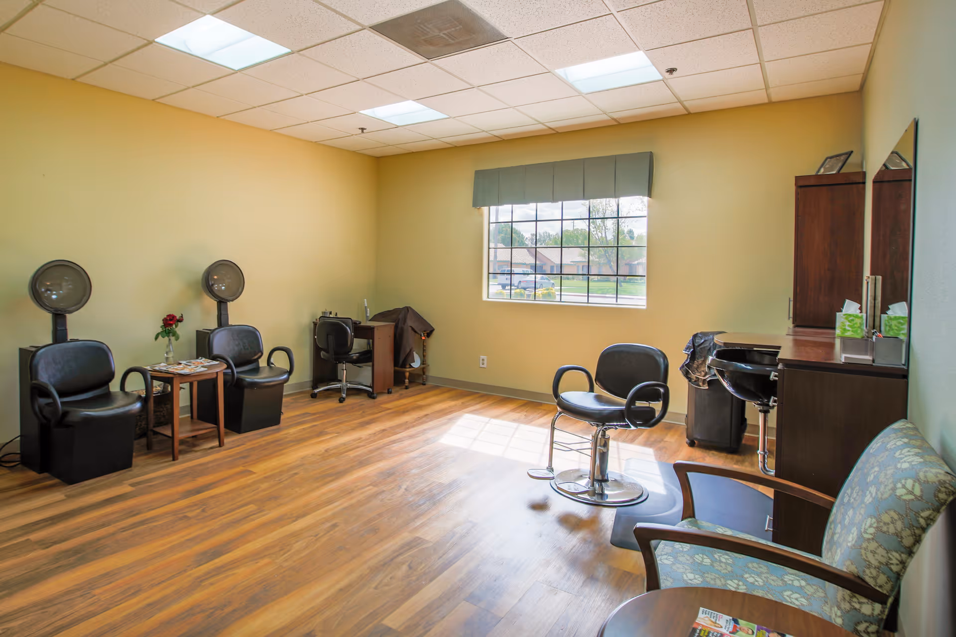 A bright room with wooden flooring and pale yellow walls, featuring two black salon hair dryer chairs with a small table between them, a black salon styling chair in front of a mirror and cabinet, a desk with a chair, and a cushioned armchair with a small round table. A window with a green valance lets in natural light.