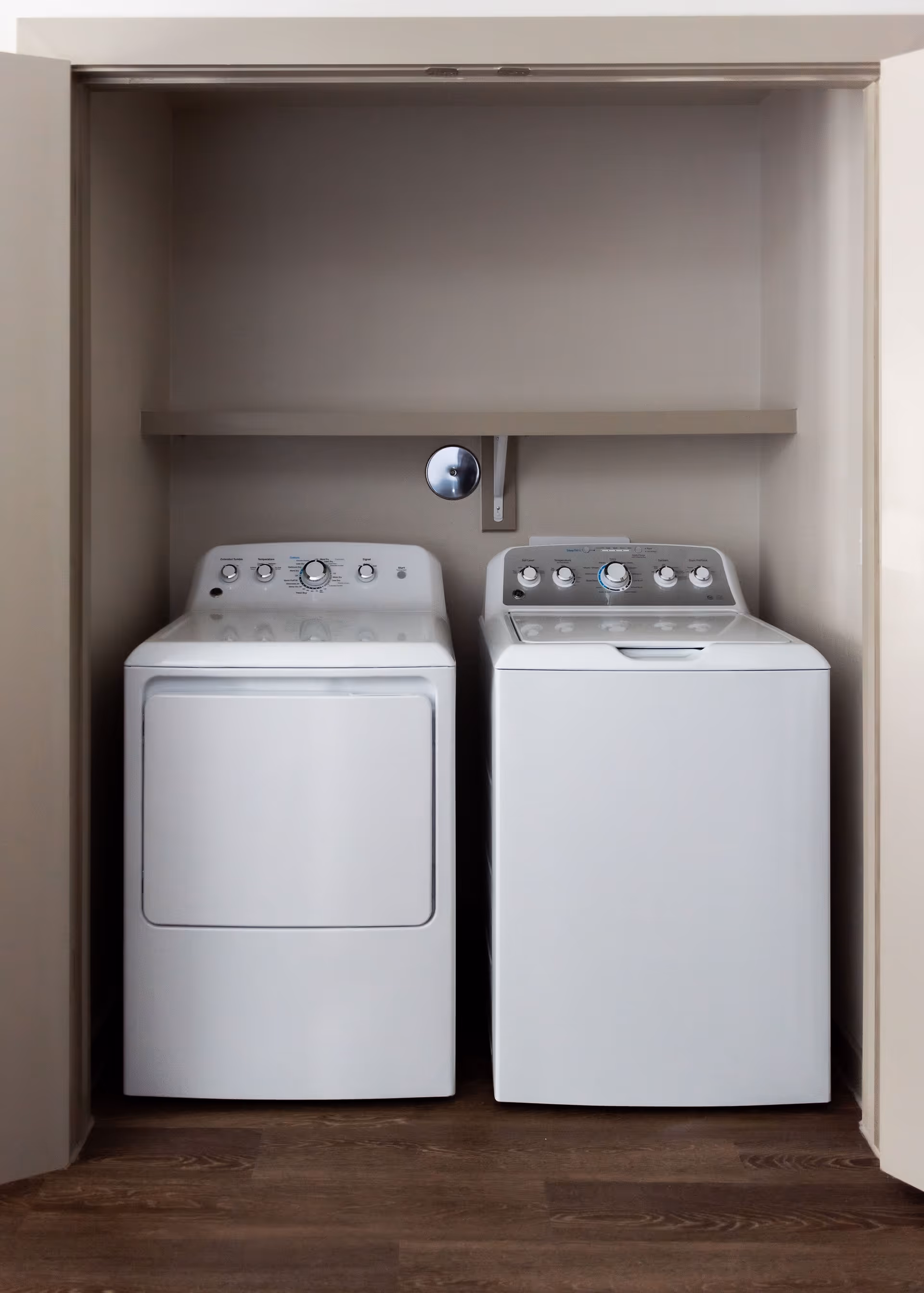 A laundry area with a white washing machine and a white dryer side by side, set inside a recessed space with beige walls and a wooden floor. There is a single shelf mounted above the machines.
