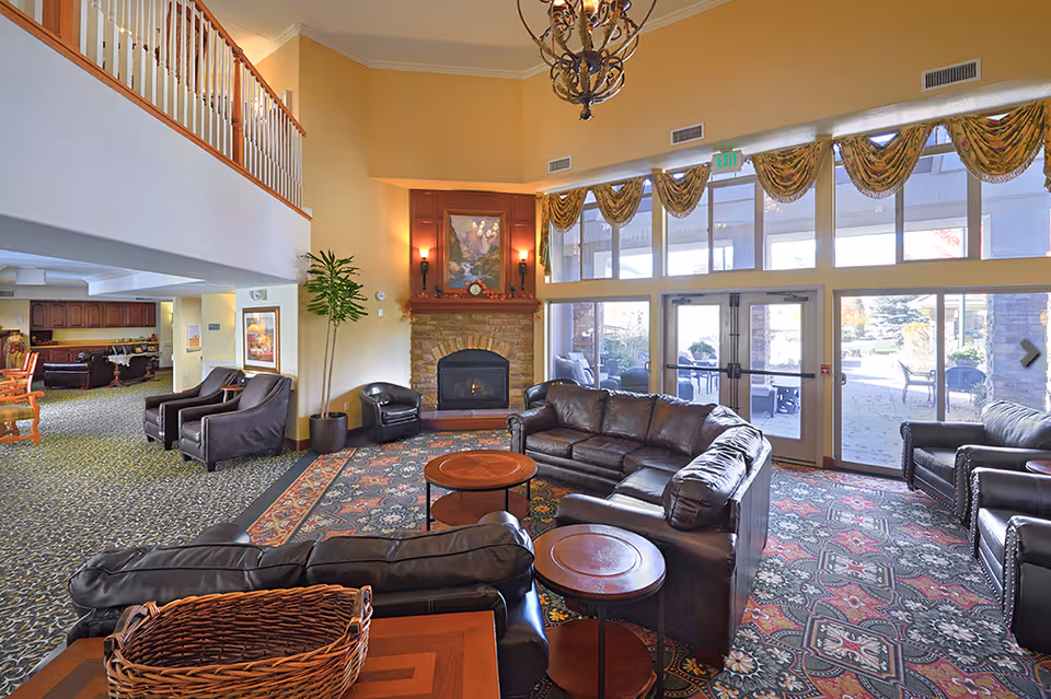 A spacious senior living facility common area with leather sofas and armchairs arranged around a stone fireplace. Large windows with decorative valances let in natural light, and a chandelier hangs from the high ceiling. There is a patterned carpet on the floor and a balcony railing visible above.