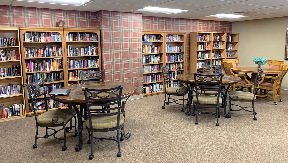 Community library/common room with round tables and chairs arranged in front of multiple bookshelves against a plaid accent wall.