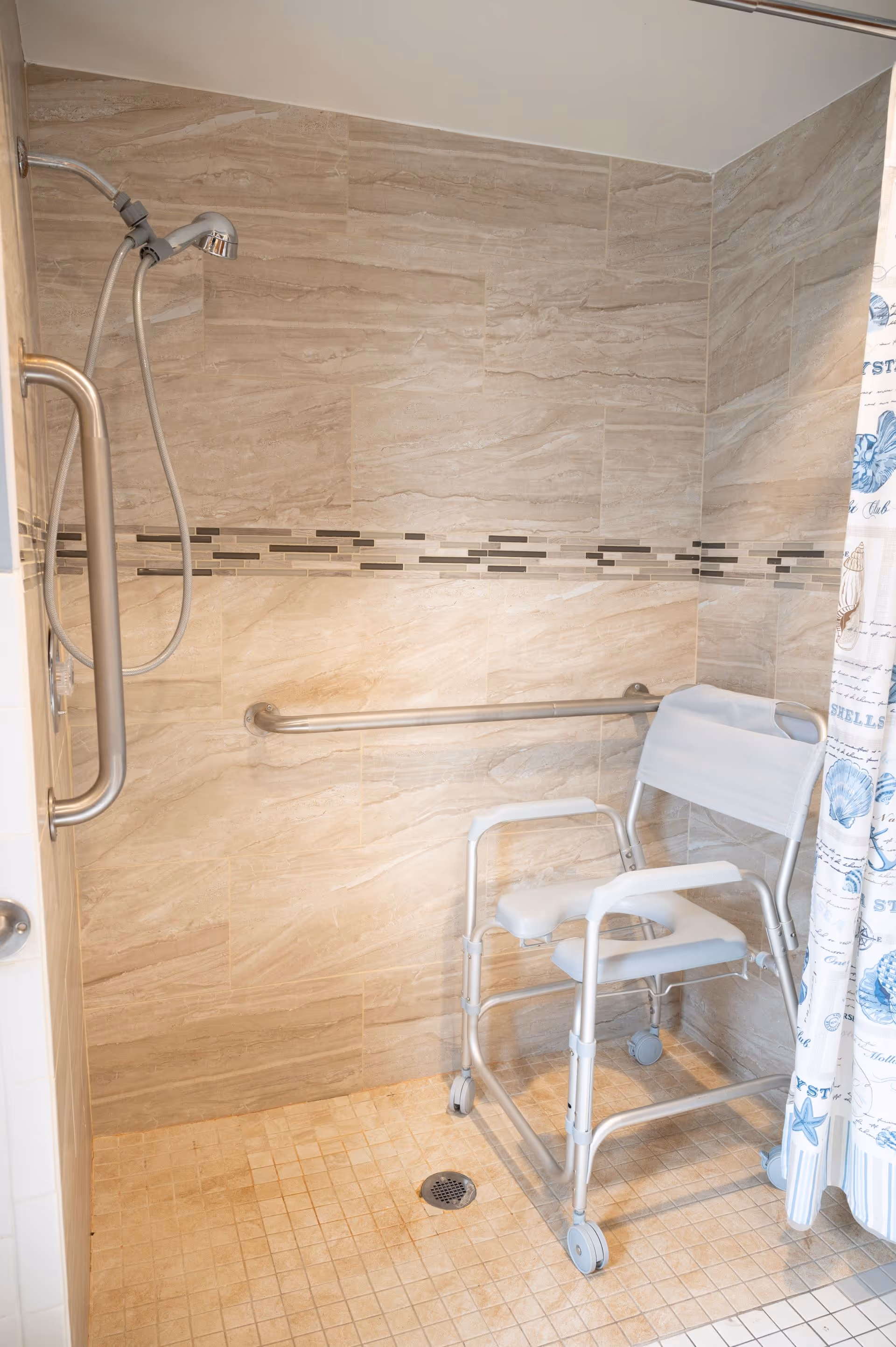 Accessible shower area with beige tiled walls and floor, a handheld showerhead, a horizontal grab bar, and a white shower chair with wheels. A shower curtain with blue seashell and starfish patterns is partially visible on the right side.