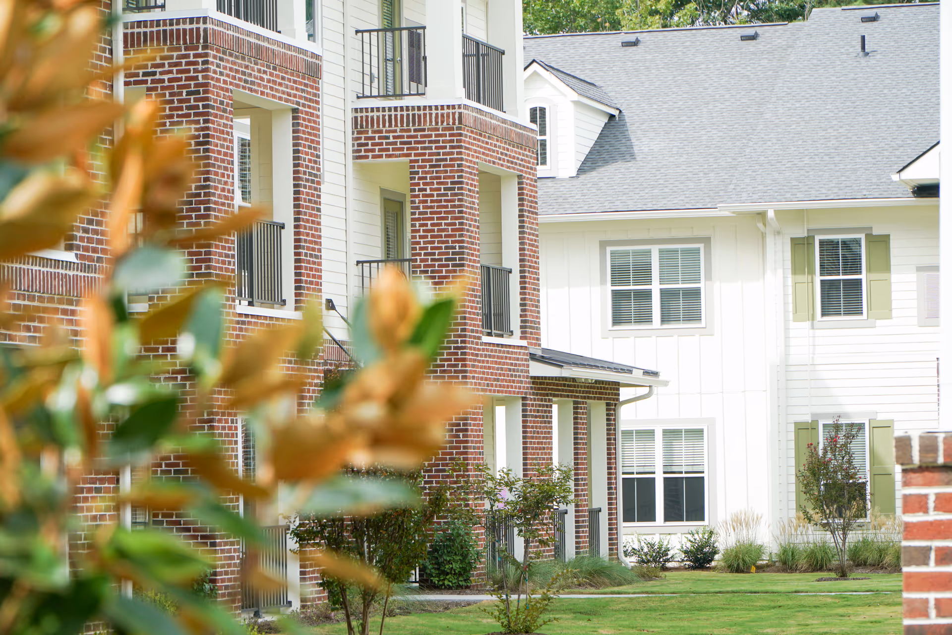 Exterior view of a red-brick and white-siding residential building with balconies and a landscaped lawn, framed by blurred foliage in the foreground.