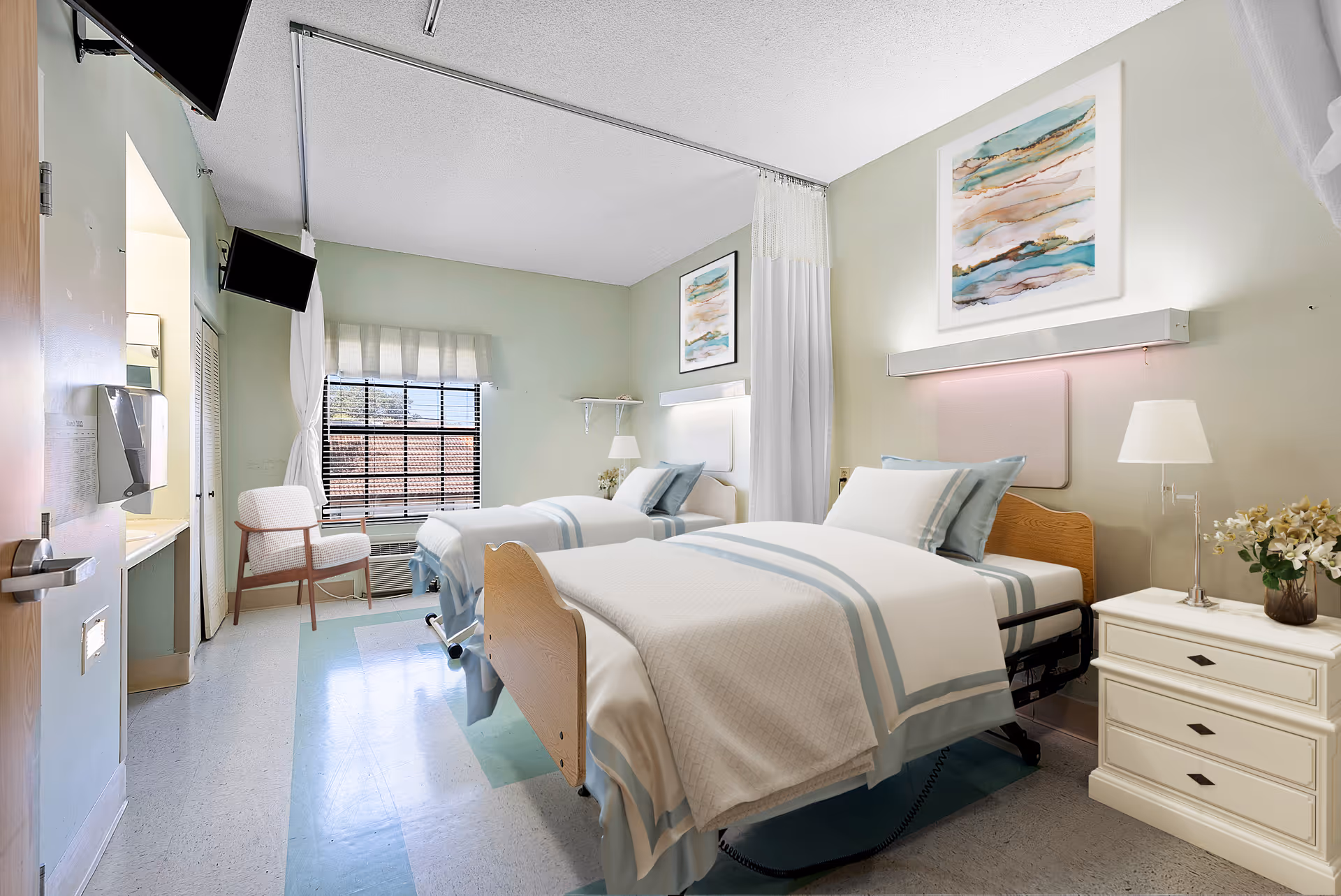 A bright and clean bedroom in a senior living facility with two single beds neatly made with white and light blue bedding. Each bed has a wooden headboard and is accompanied by a white nightstand with a lamp and a vase of flowers. There are two framed abstract paintings on the walls above the beds. A window with blinds and a white chair are visible at the far end of the room. The room has light green walls and a light-colored floor with a blue rug under the beds. A TV is mounted on the wall near the entrance door.