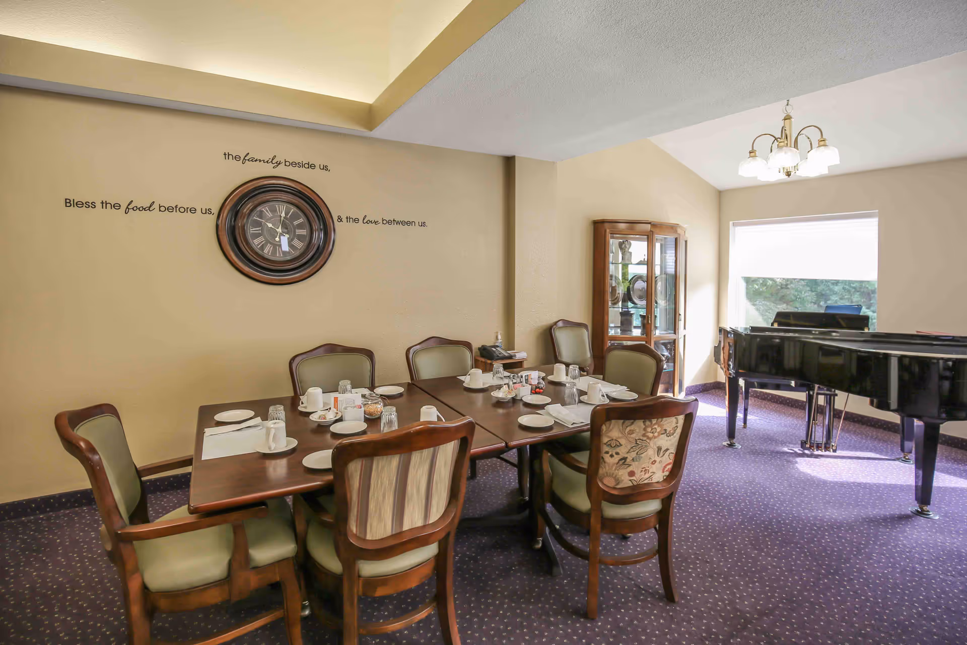 A dining room with a set table surrounded by chairs, a wall clock and decorative lettering, and a black grand piano by the window.