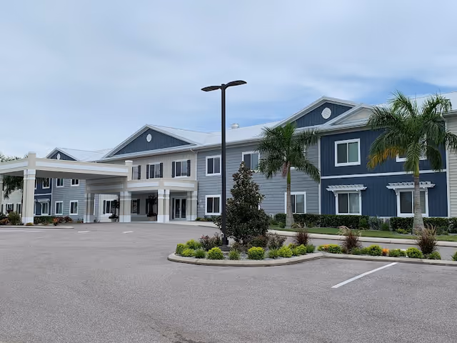 Front entrance of a two-story coastal-style senior living building with a covered porte-cochere, palm trees, and an empty parking area.