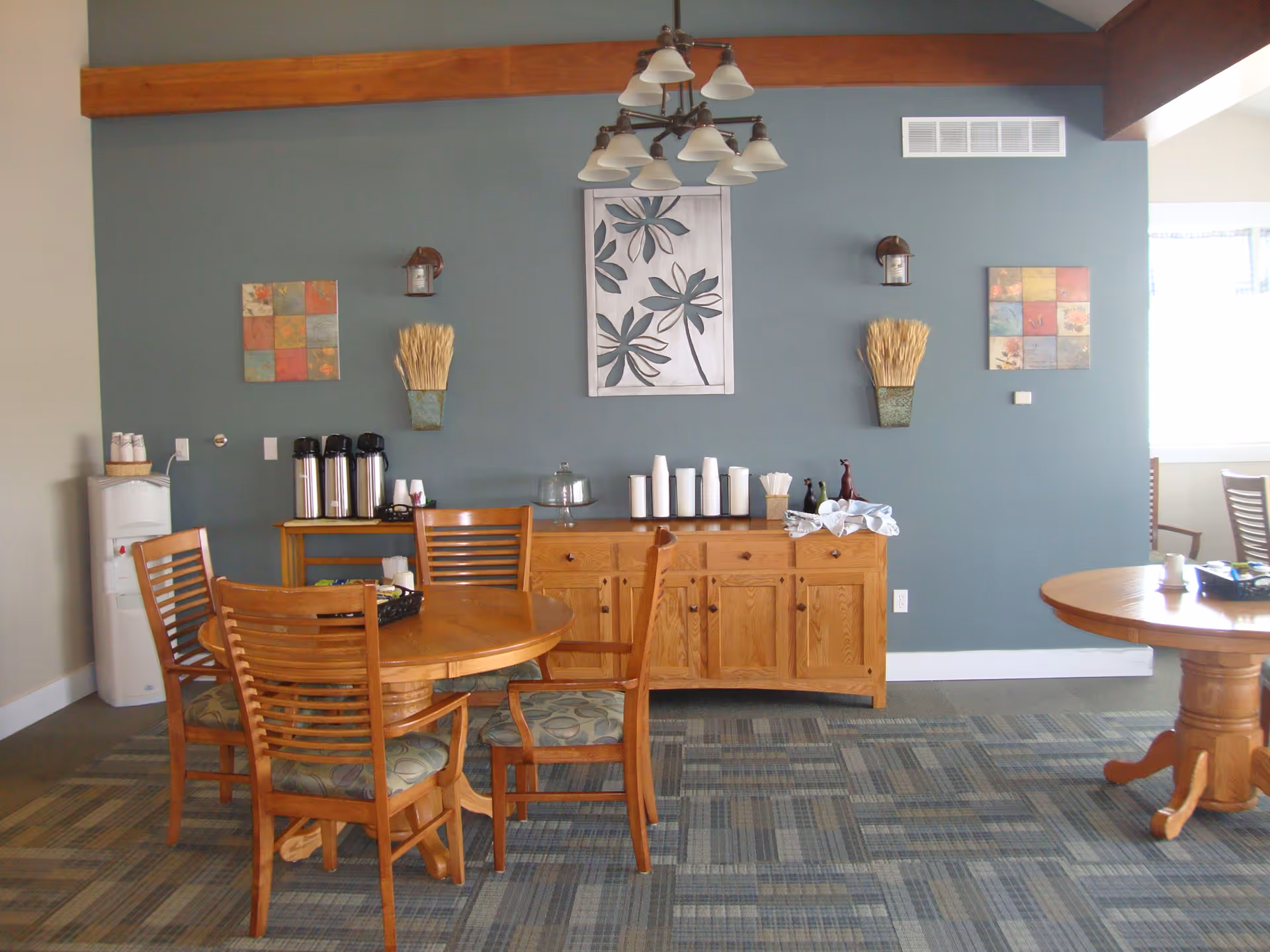 A cozy dining area with wooden tables and chairs, a sideboard with coffee dispensers, cups, and condiments, decorative wall art, and a ceiling light fixture. The walls are painted blue and beige, and the floor is carpeted with a patterned design.