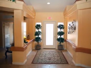 Interior hallway with beige walls and tiled floor leading to a white door with an exit sign above it. Two potted plants with green foliage are placed on either side of the door. A patterned rug is centered on the floor. The hallway has built-in ledges and decorative columns.
